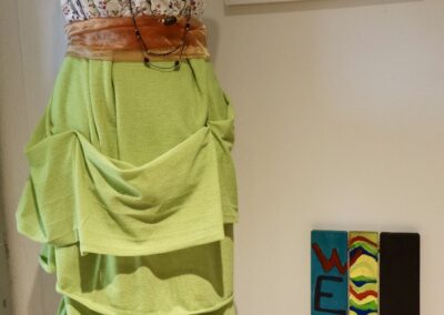 Dekorative Schneiderpuppe mit asymmetrischem Kleid aus grünem Stoff und geblümtem Oberteil vor einem Schild mit Öffnungszeiten