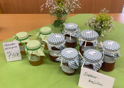 Gläser mit Gelee und Konfitüre in karierten Stoffdeckeln auf grünem Tuch, mit Preisschildern und Blumenvasen