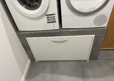Weißes Podest mit Schublade unter Waschmaschine und Trockner auf grauen Fliesen